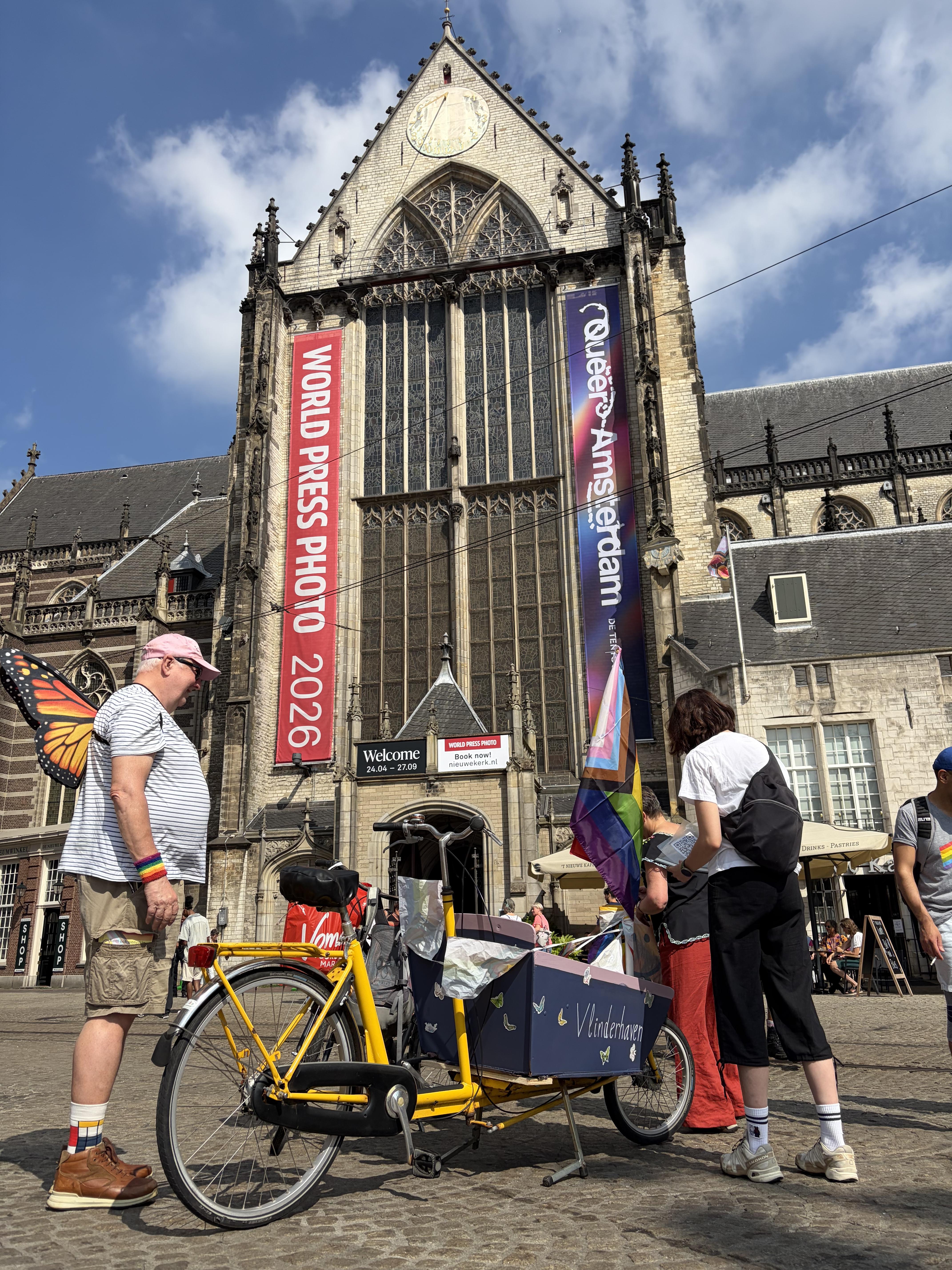 The New Church in Amsterdam with a massive World Press Photo 2026 banner in the front. some people and a hauling bicycle with a trans pride flag in front on the street