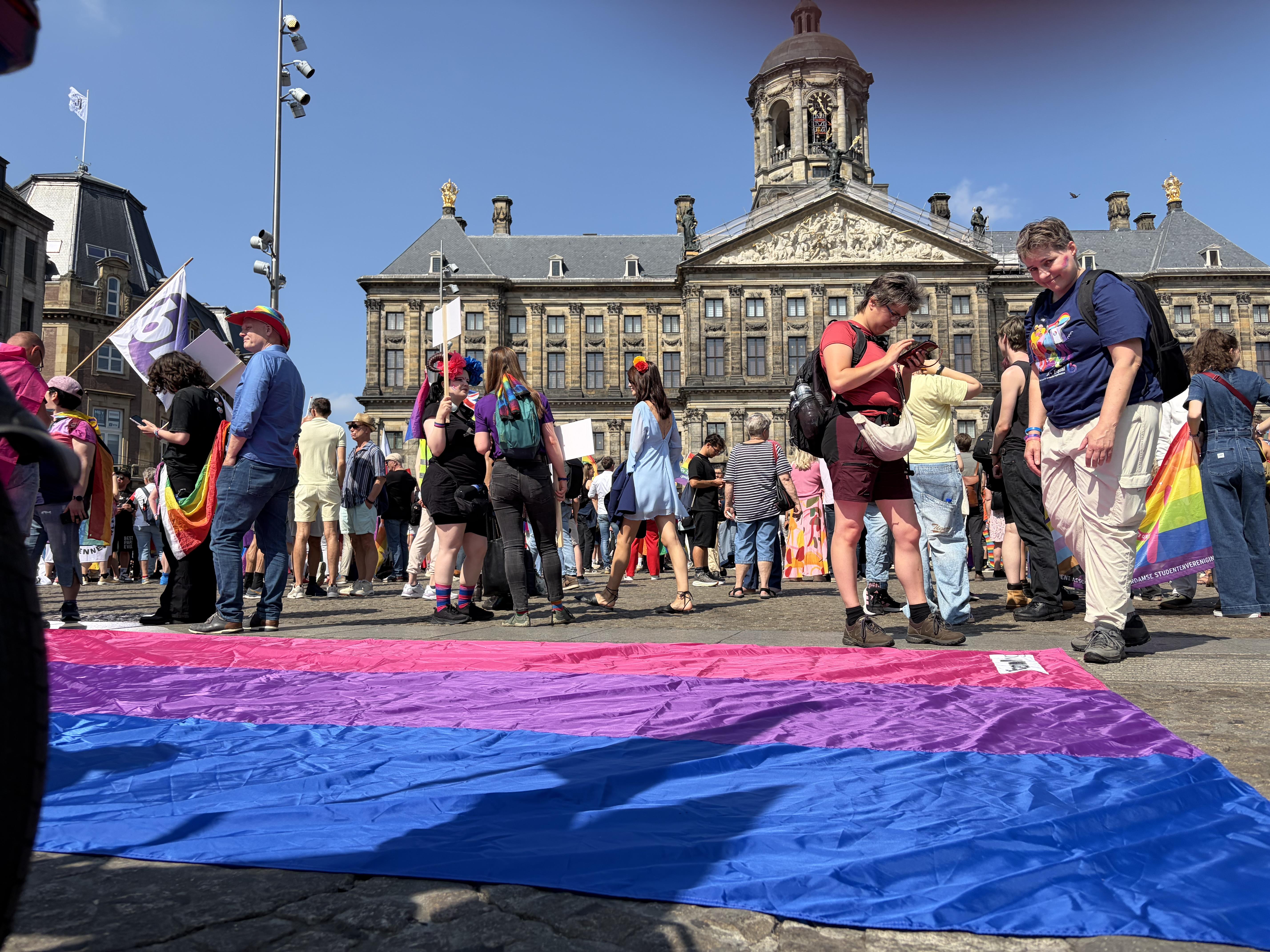 Bi Pride flag lays on the ground in Dams Square Amsterdam with many people walking and standing in the background in front of Royal Palace Amsterdam
