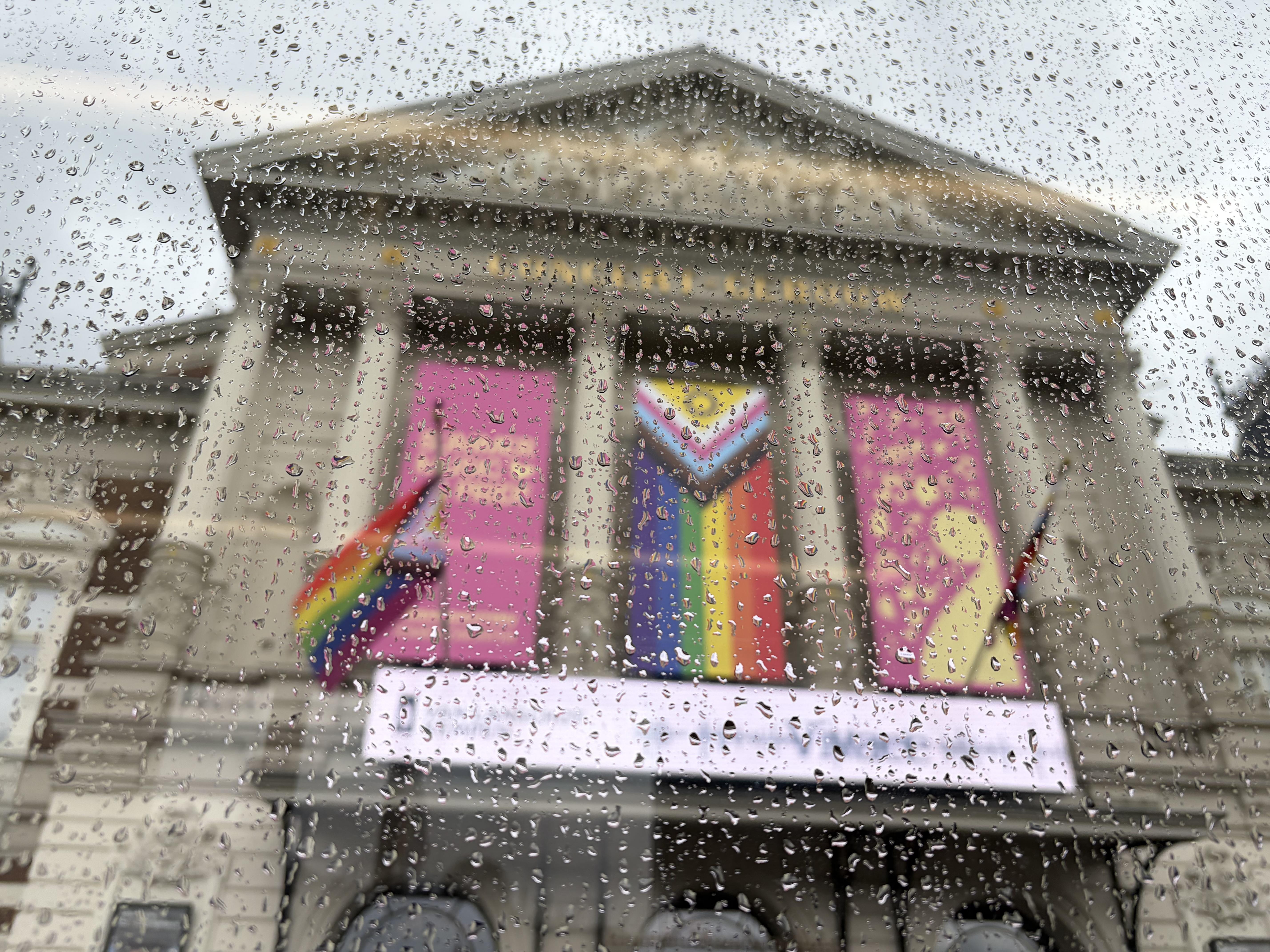 Het Concertgebouw in Amsterdam through the window of a tram with raindrops on the window in focus, trans pride flags wave in the background