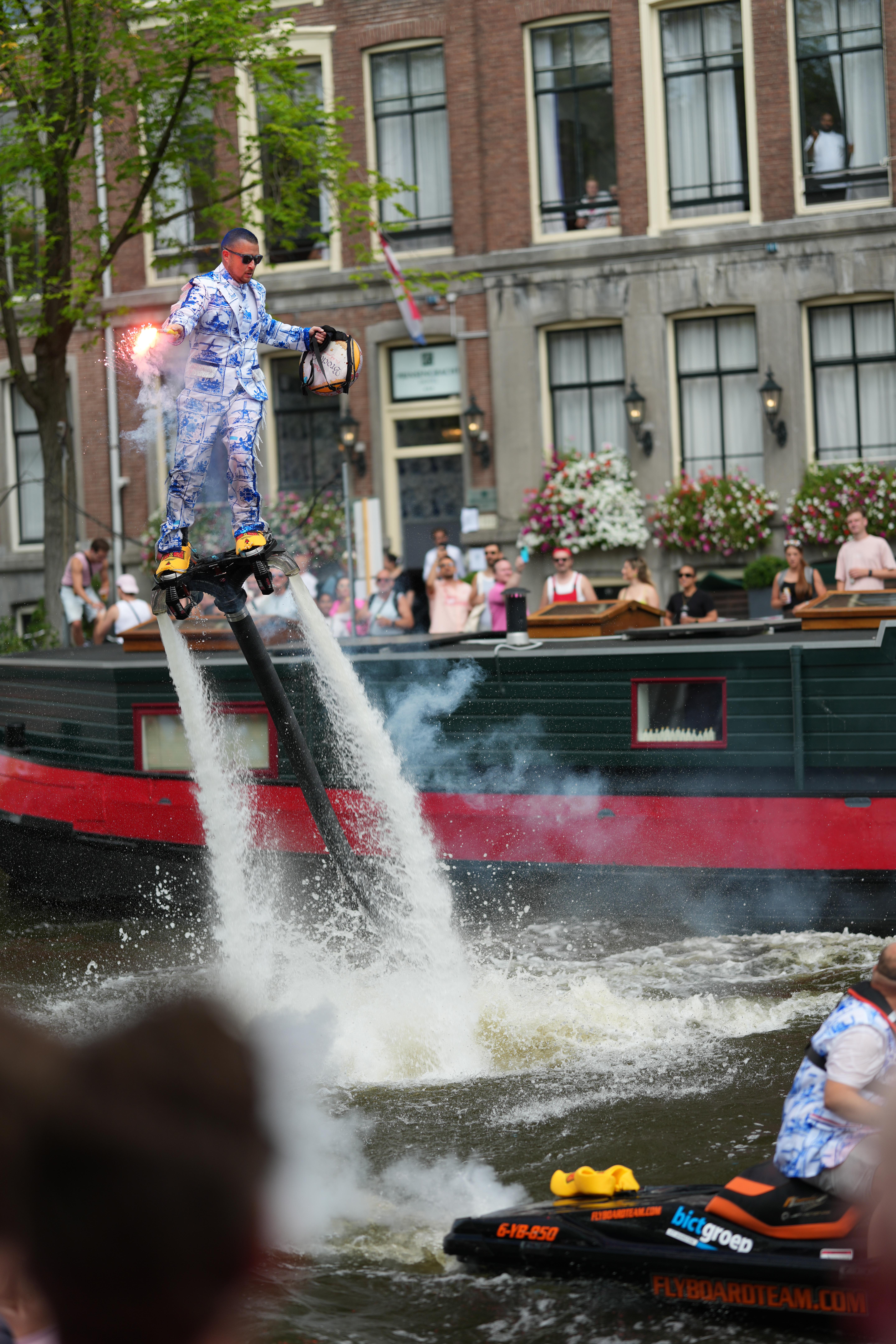 a guy wearing stereotypical Dutch things, porcelain doll looking clothing and clogs, while flying around with water jet shoes at Canal Pride Amsterdam, preparing to do a flip in the air