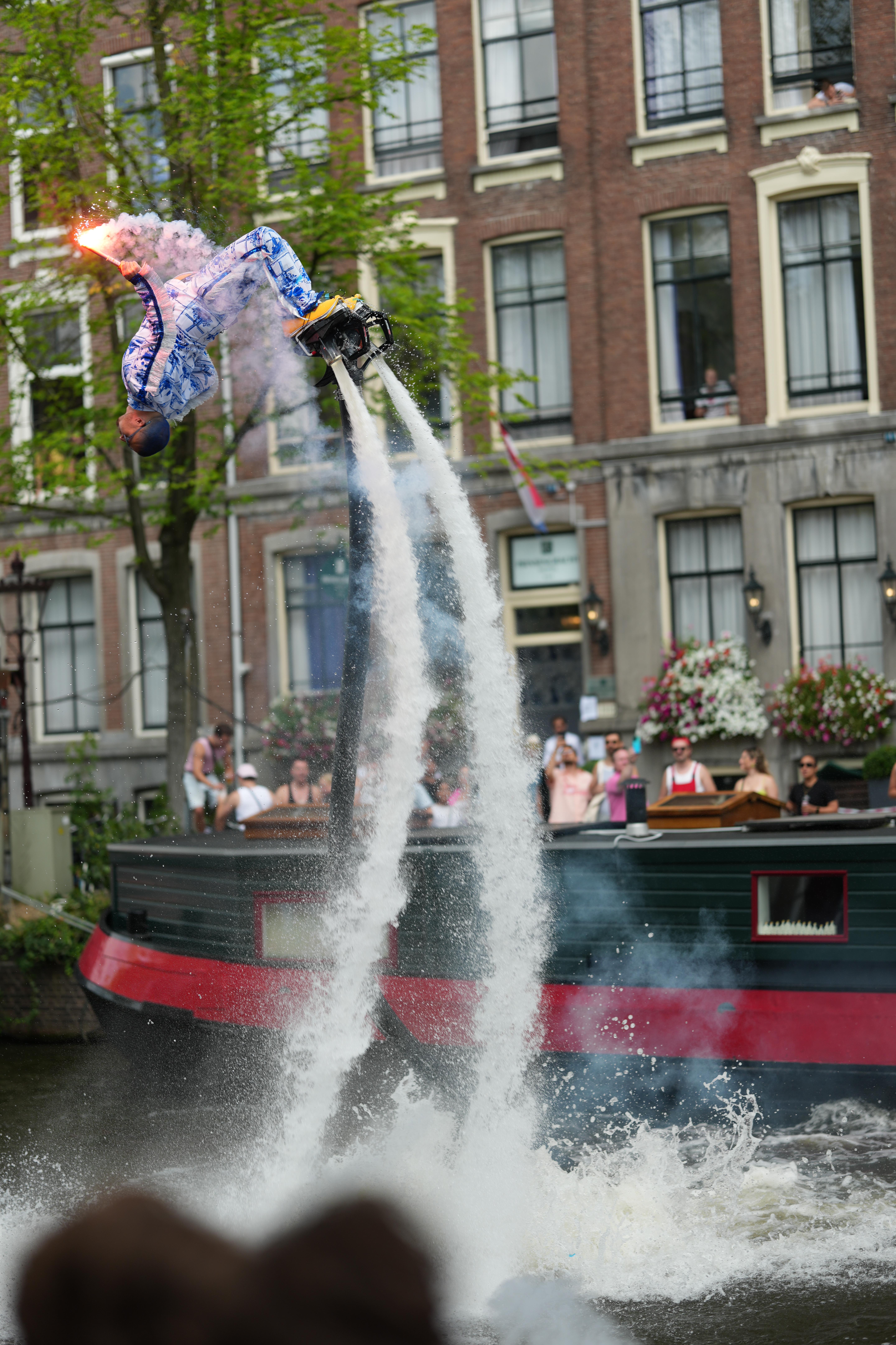 a guy wearing stereotypical Dutch things, porcelain doll looking clothing and clogs, while flying around with water jet shoes at Canal Pride Amsterdam, performing a flip in the air