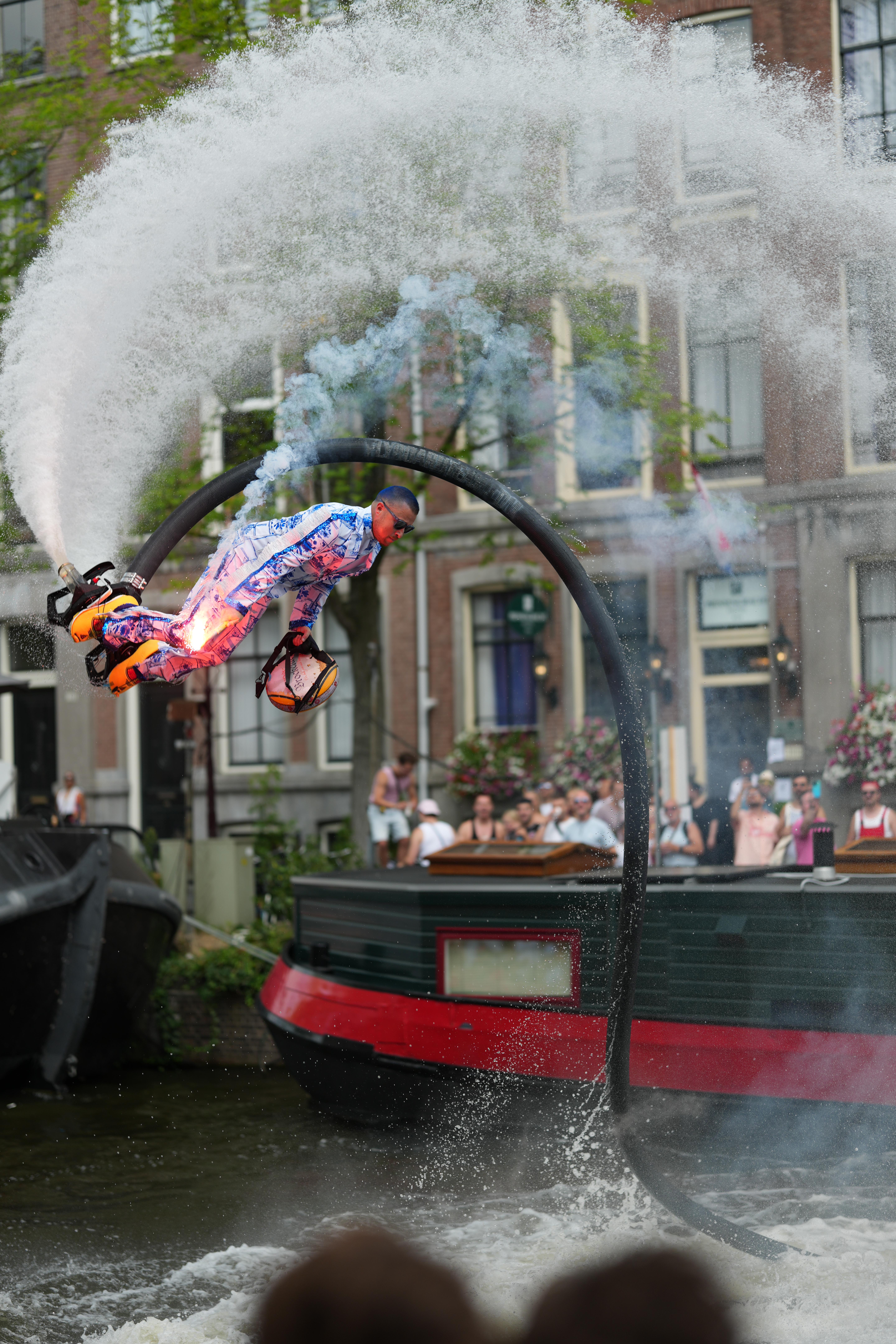 a guy wearing stereotypical Dutch things, porcelain doll looking clothing and clogs, while flying around with water jet shoes at Canal Pride Amsterdam, performing a flip in the air