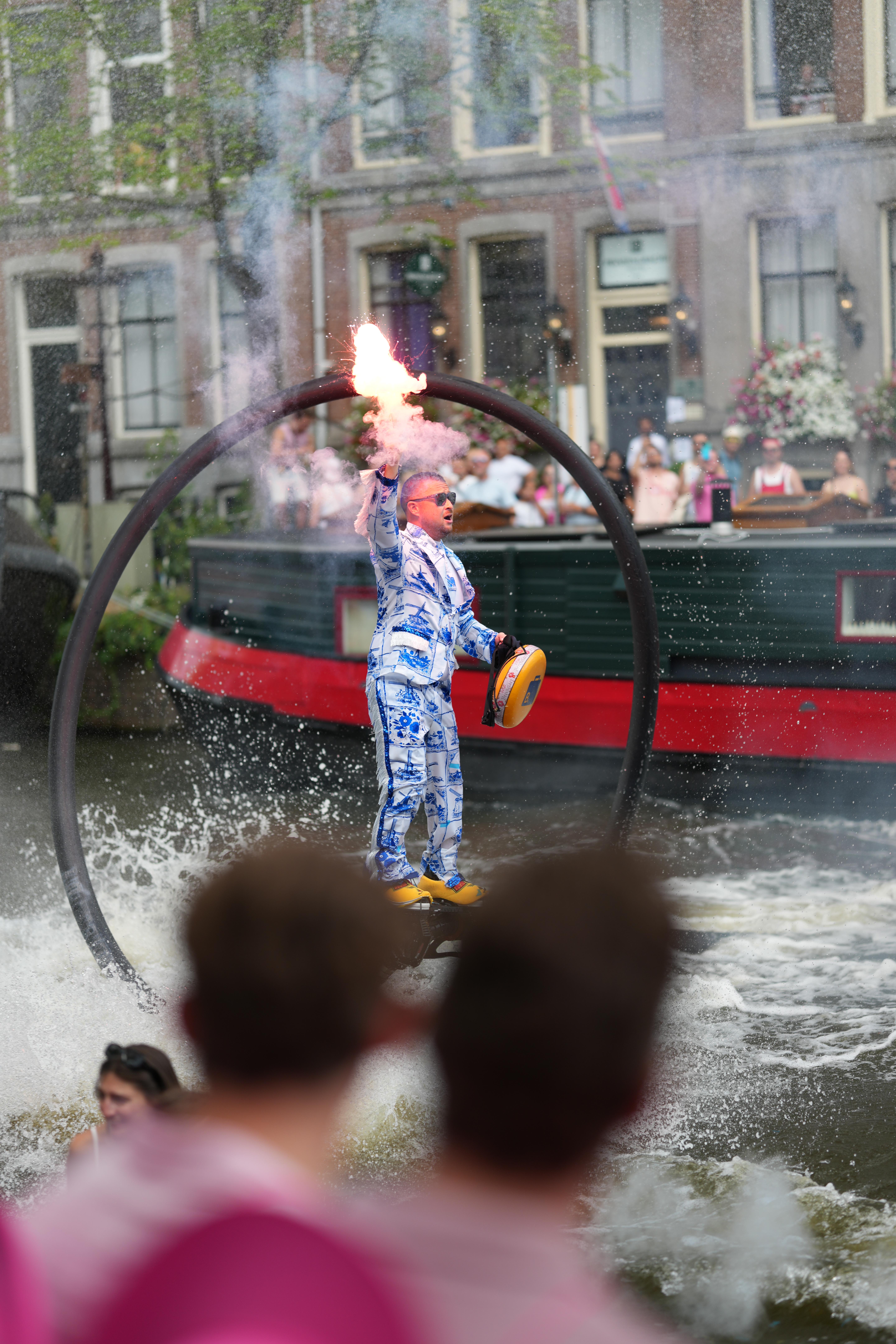 a guy wearing stereotypical Dutch things, porcelain doll looking clothing and clogs, while flying around with water jet shoes at Canal Pride Amsterdam, performing a flip in the air