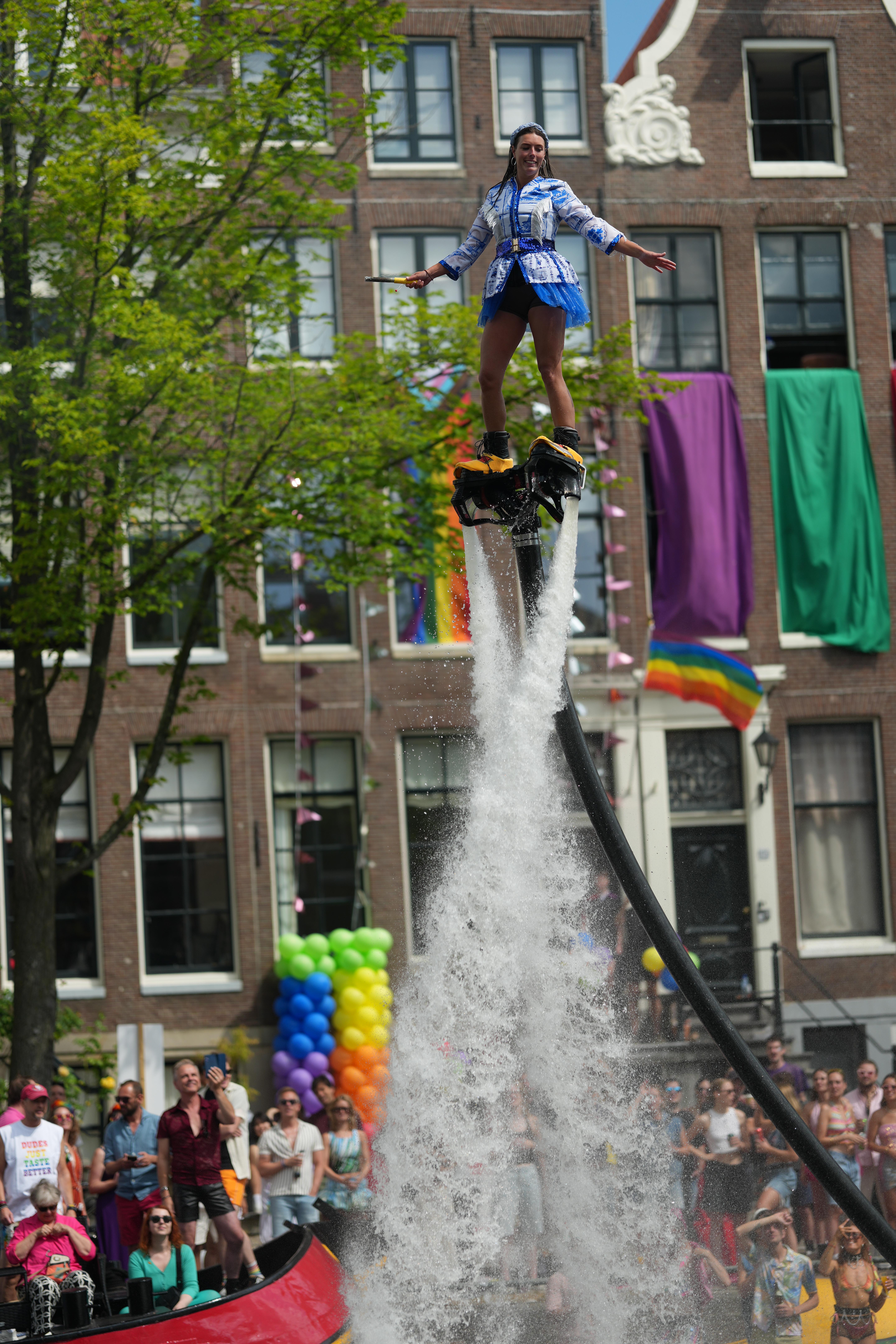 a Dutch woman wearing water jet shoes flying in the air and wearing stereotypical Dutch things, porcelain doll dress and clogs. people stand and sit on the street in the background, at Canal Pride Amsterdam