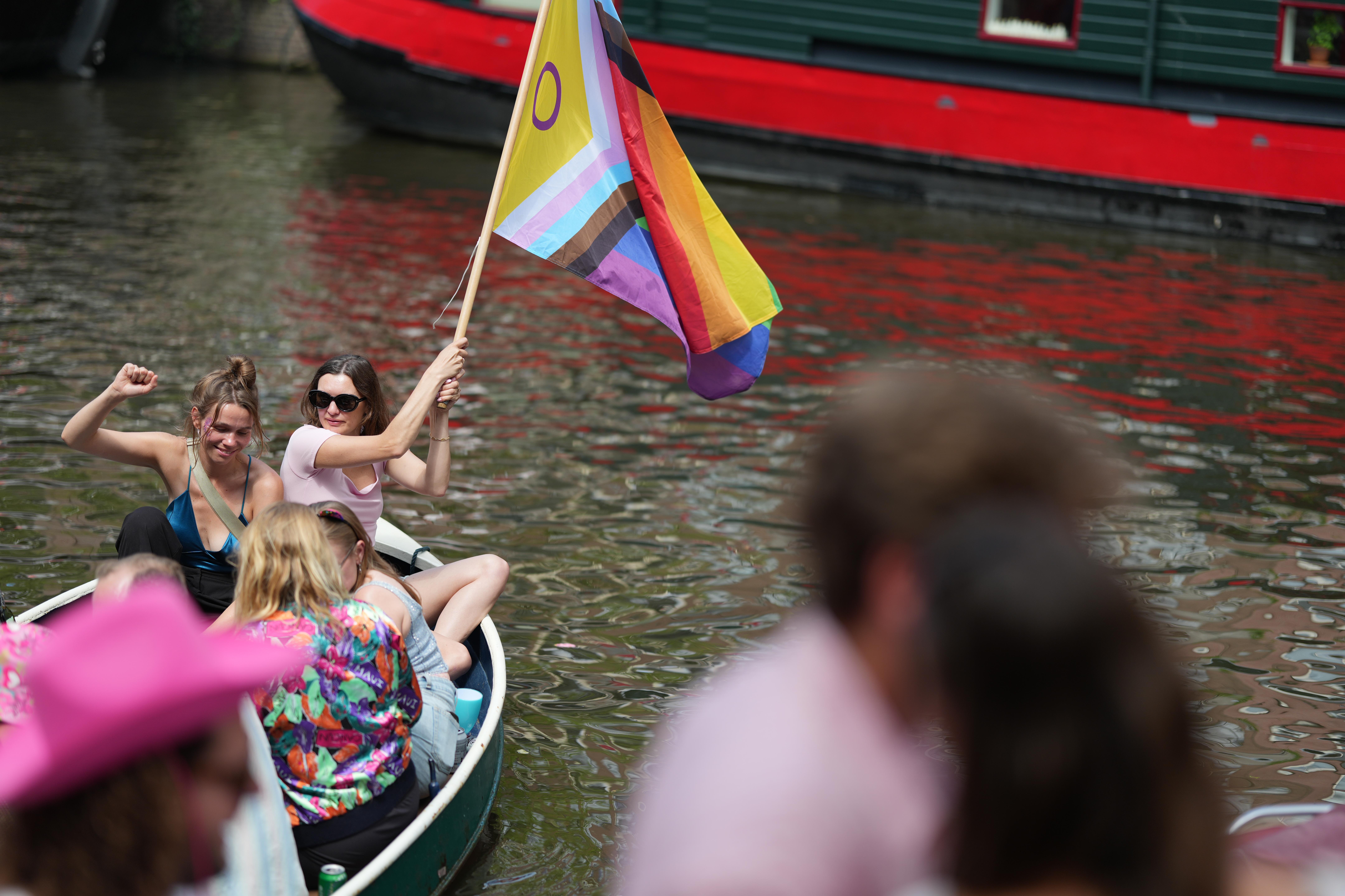 a person and their friends waving a pride flag in a small boat at Canal Pride in Amsterdam, with some blurred out people in the foreground