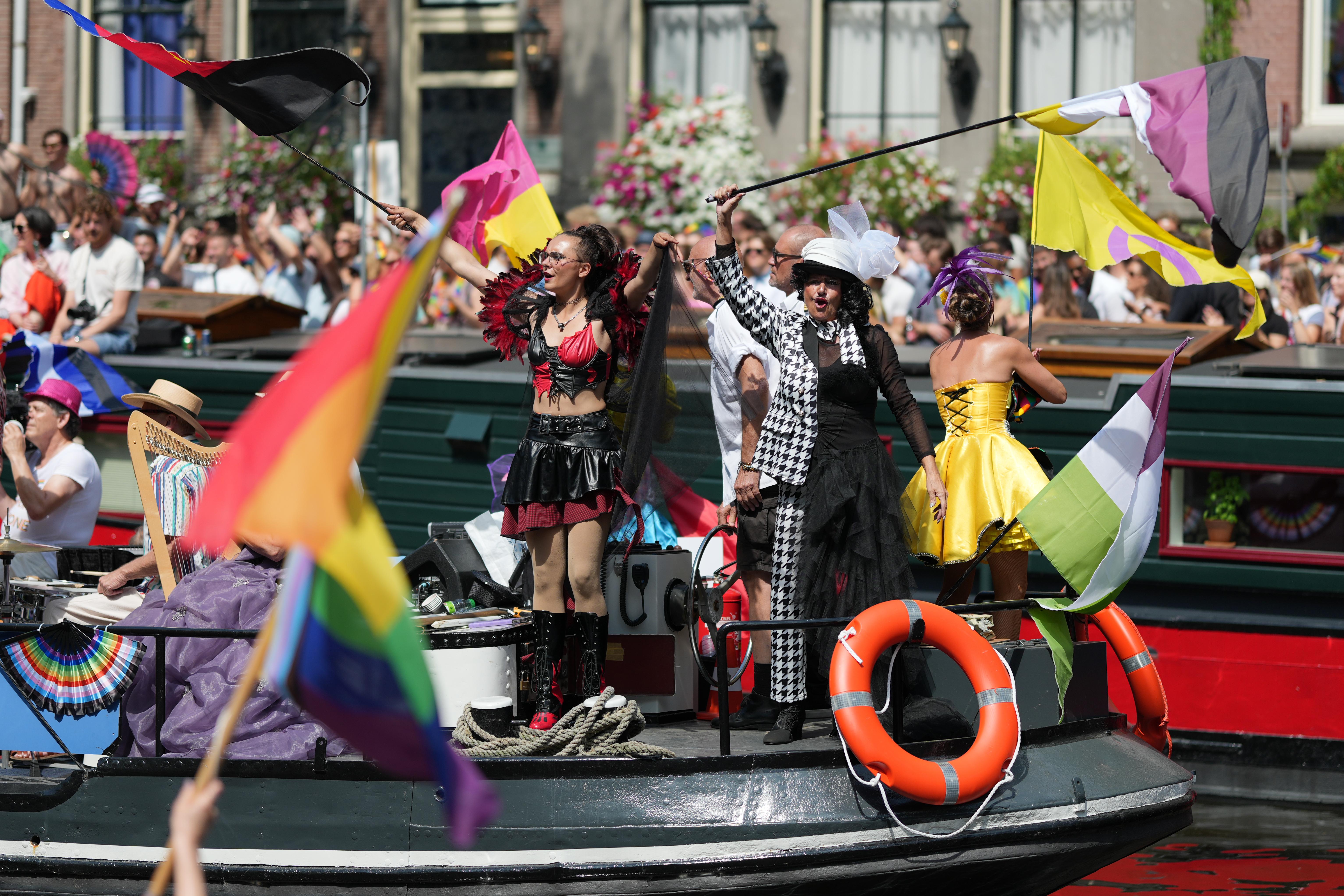 two people amazingly dressed folx in focus waving two pride flags, with many other pride flags blurred out in the foreground and background, at Canal Pride in Amsterdam