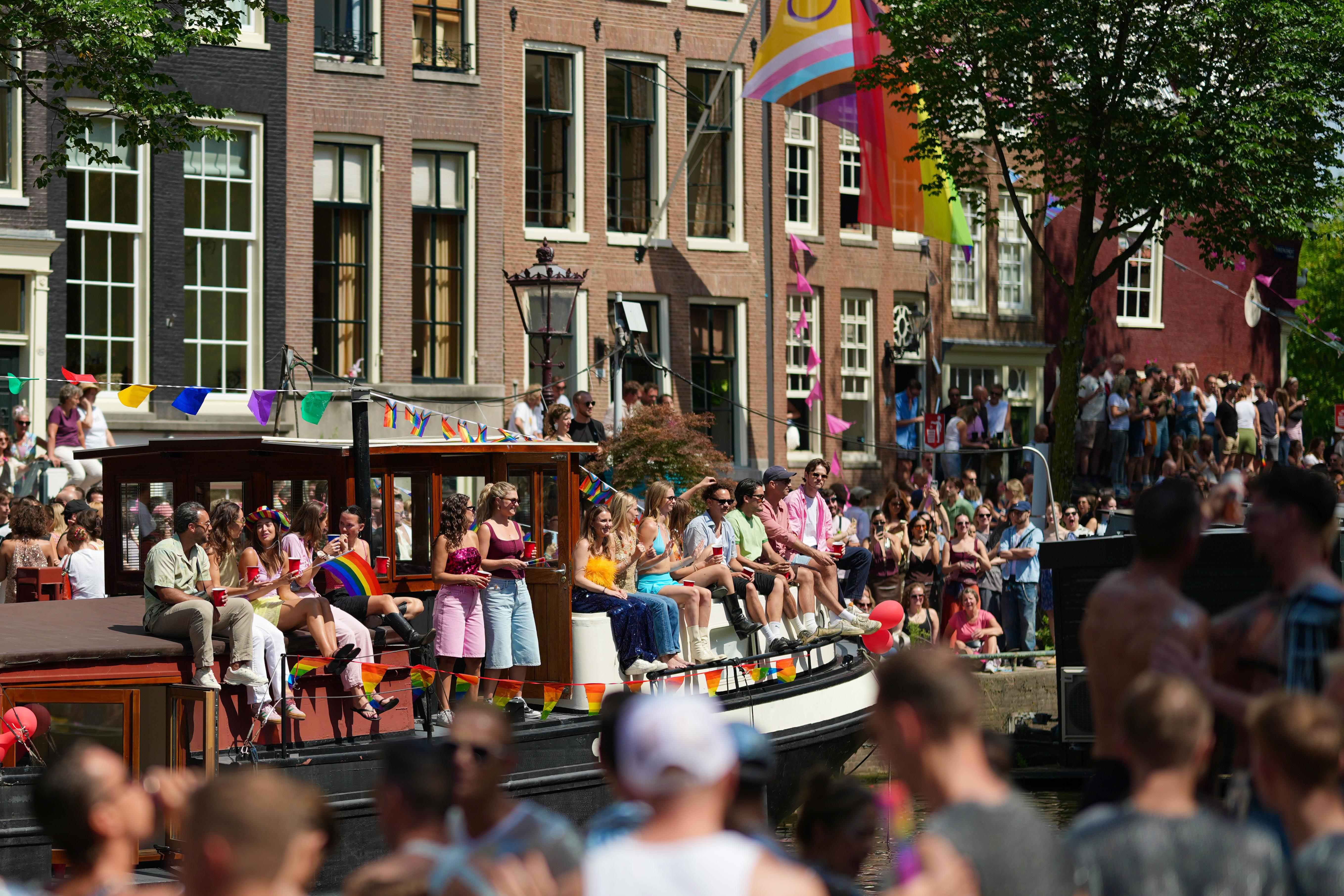 many people sitting in a row on a house boat on the canal during Canal Pride in Amsterdam. many people blurred out in the foreground with a giant progressive pride flag waving in the background