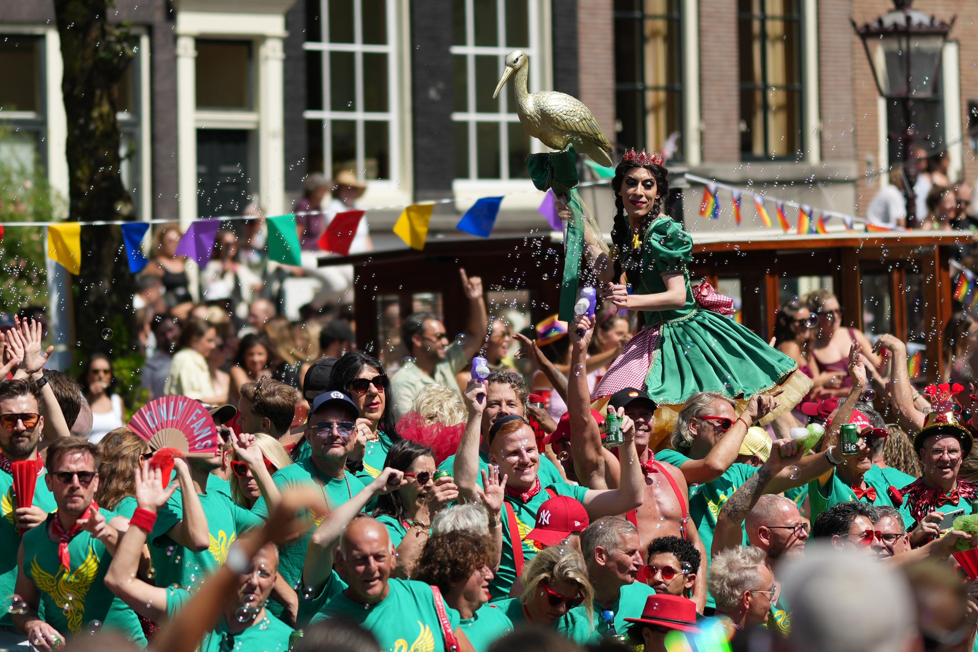 many people dance, wave fans, and blow bubbles from bubble guns on a boat at Canal Pride in Amsterdam. one person wearing a crown is in focus holds a golden crane